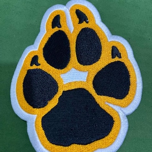 Puede incluir: Un parche bordado con forma de huella de pata que presenta una almohadilla central grande y cuatro almohadillas pequeñas en hilo negro. El diseño está delineado con hilo amarillo vibrante y tiene un borde blanco grueso en todo el contorno.