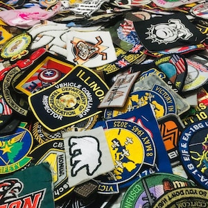 May include: A collection of embroidered patches in various shapes and colors. Many patches feature text, logos, and emblems, including military, vehicle inspection, and security service designs. The patches are densely packed together.