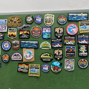 May include: A collection of various embroidered patches featuring mountain landscapes, outdoor activities, and travel destinations pinned to a green board. The patches display diverse shapes, colors, and text including names like Mt Princeton, La Thuile, and Livigno.