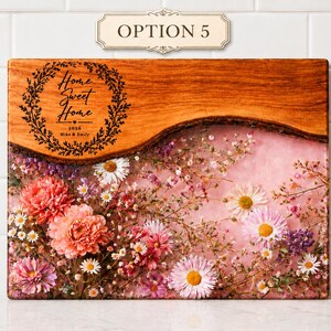 May include: Rectangular wooden cutting board with a live edge and a floral resin design. The top section is engraved with "Home Sweet Home" and the names "Mike & Emily" and the year 2026. The resin section is filled with pink and white flowers.