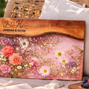 May include: A rectangular wooden serving board with a live edge, featuring a pink resin inlay filled with colorful flowers. The top edge has the names "JORDAN & KEVIN" and a heart symbol. A decorative piece for serving.