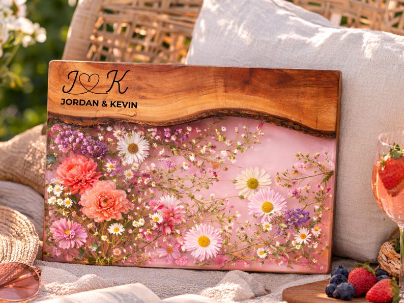 May include: A rectangular wooden serving board with a live edge, featuring a pink resin inlay filled with colorful flowers. The top edge has the names "JORDAN & KEVIN" and a heart symbol. A decorative piece for serving.