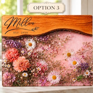 May include: A rectangular wooden cutting board with a live edge, featuring a floral design with pink, purple, and white flowers encased in resin. The top section has the word "Miller" and "EST. 2026" in black script.