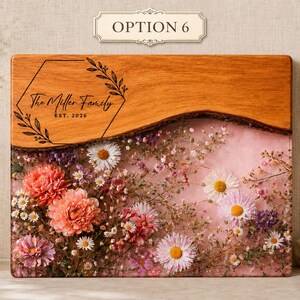 May include: A rectangular wooden cutting board with a floral resin design. The top section is natural wood, while the bottom features a pink resin with embedded flowers. The text "The Miller Family EST. 2026" is engraved on the wood.