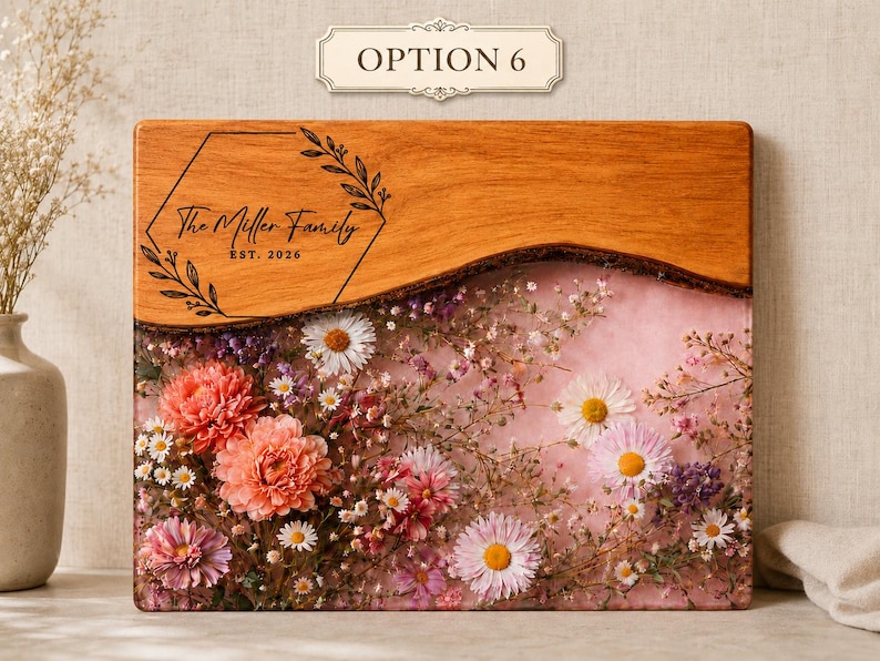 May include: A rectangular wooden cutting board with a floral resin design. The top section is natural wood, while the bottom features a pink resin with embedded flowers. The text "The Miller Family EST. 2026" is engraved on the wood.