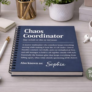 May include: A navy blue spiral-bound notebook with the words "Chaos Coordinator" in white. The definition of the term is printed below, along with the name "Sophia". A gold and white pen and a pair of wireless earbuds are also visible.