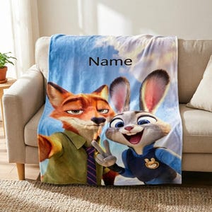 May include: A plush blanket featuring the animated characters Nick Wilde and Judy Hopps from Zootopia. The blanket has a blue sky background with the word "Name" printed on it. The characters are in full color, with Nick in a green shirt and Judy in a blue uniform.