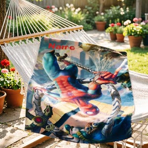May include: A rectangular blanket featuring a vibrant graphic of a superhero in a red and blue suit suspended upside down, interacting with mechanical tentacles. The word Name is printed in red at the top of the fabric, which is draped over a white hammock.