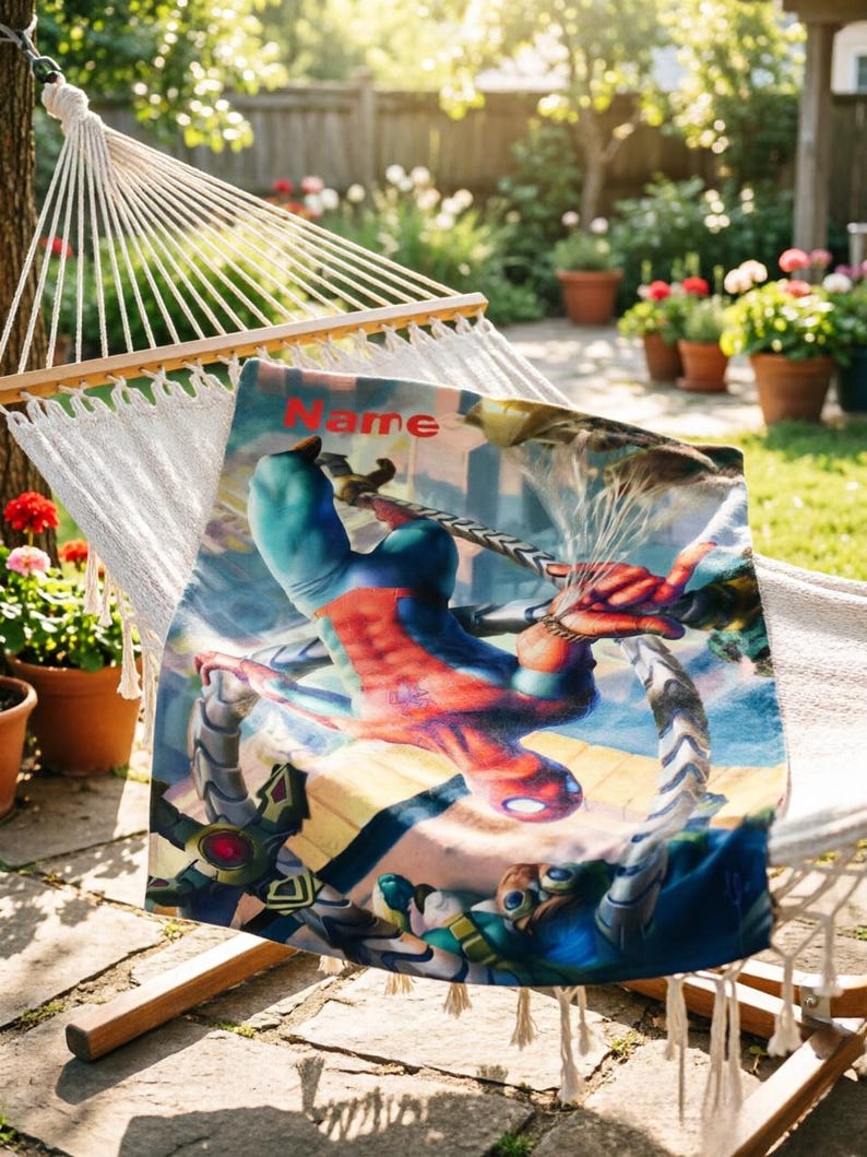 May include: A rectangular blanket featuring a vibrant graphic of a superhero in a red and blue suit suspended upside down, interacting with mechanical tentacles. The word Name is printed in red at the top of the fabric, which is draped over a white hammock.