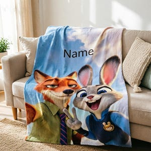 May include: A plush blanket featuring the animated characters Nick Wilde and Judy Hopps from Zootopia. The blanket has a blue sky background with the word "Name" printed on it. The characters are in a selfie pose.