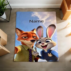 May include: A rectangular blanket featuring the characters Nick Wilde and Judy Hopps from Zootopia, set against a blue sky background. The blanket has the word "Name" printed on it. The fox is wearing a green shirt and the bunny is wearing a blue uniform.