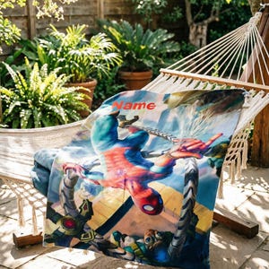 May include: A rectangular blanket featuring a colorful graphic print of a superhero in a red and blue suit suspended by mechanical tentacles. The word Name is printed in orange letters at the top of the fabric, which displays a dynamic scene.