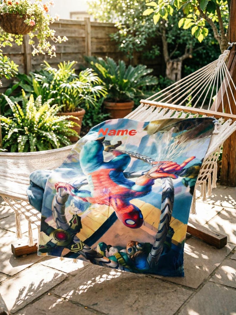 May include: A rectangular blanket featuring a colorful graphic print of a superhero in a red and blue suit suspended by mechanical tentacles. The word Name is printed in orange letters at the top of the fabric, which displays a dynamic scene.