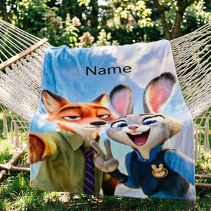May include: A blue blanket featuring Nick Wilde and Judy Hopps from Zootopia, with the word "Name" printed on it. The blanket is draped over a hammock in a garden setting. The characters are in a selfie pose.