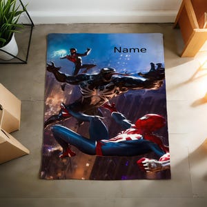 May include: A rectangular blanket featuring a graphic print of Spider-Man and Venom in a dynamic battle scene against a dark city background. The design includes a smaller figure in the upper left and the word Name printed in black text.