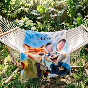 May include: A blue and white blanket featuring cartoon characters Nick Wilde and Judy Hopps from Zootopia. The blanket has the word "Name" printed on it. The blanket is draped over a hammock in a garden setting.
