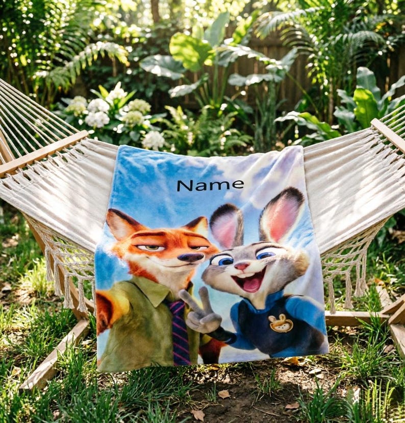 May include: A blue and white blanket featuring cartoon characters Nick Wilde and Judy Hopps from Zootopia. The blanket has the word "Name" printed on it. The blanket is draped over a hammock in a garden setting.