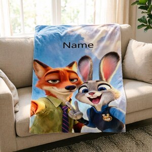 May include: A plush blanket featuring the characters Nick Wilde and Judy Hopps from Zootopia, set against a sky background. The blanket has the word "Name" printed on it. The blanket is draped over a beige sofa.