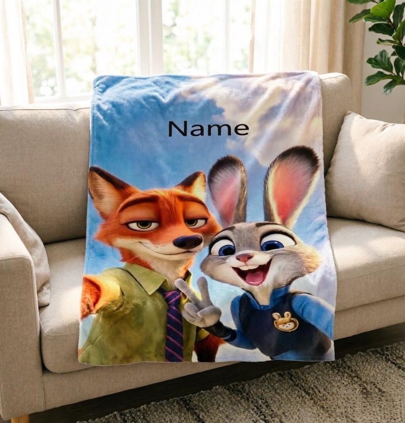 May include: A plush blanket featuring the characters Nick Wilde and Judy Hopps from Zootopia, set against a sky background. The blanket has the word "Name" printed on it. The blanket is draped over a beige sofa.
