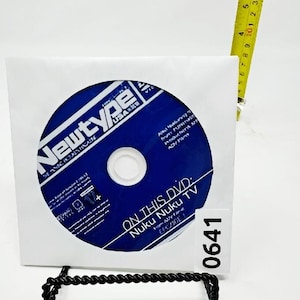 Puede incluir: Un disco DVD azul en una funda de papel blanca, con el texto "Newtype" y "ON THIS DVD: Nuku Nuku TV" visibles. Una cinta métrica amarilla está al lado del disco. El disco está sujeto por un soporte de metal negro. También hay una pegatina blanca con el número "0641".