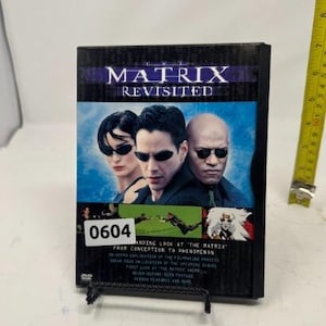 Könnte beinhalten: DVD-Hülle für "The Matrix Revisited" mit Bildern der Filmstars. Die Hülle ist schwarz mit einem lila-blauen Banner. Die DVD wird auf einem schwarzen Metallständer präsentiert. Die Hülle ist etwa 19 cm hoch.