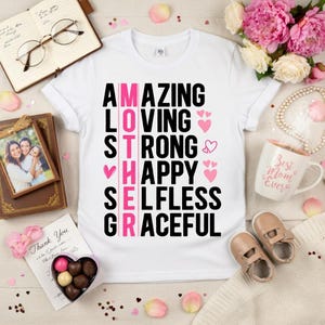 May include: White t-shirt with the word "MOTHER" in pink and black text, with the words "AMAZING, LOVING, STRONG, HAPPY, SELFLESS, GRACEFUL". The shirt is surrounded by flowers, a mug, and baby shoes.