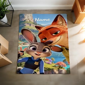 May include: A rectangular blanket featuring the animated characters Judy Hopps and Nick Wilde from Zootopia. Judy, a gray rabbit, is in a police uniform, giving a peace sign. Nick, an orange fox, smiles. The word "Name" is at the top.