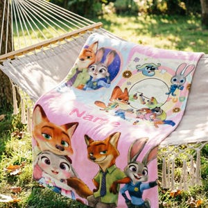 May include: A soft pink fleece blanket featuring colorful cartoon illustrations of a fox and a rabbit character from Zootopia. The design includes multiple scenes of the characters, a circular frame, and the word Name printed in a bold pink font.