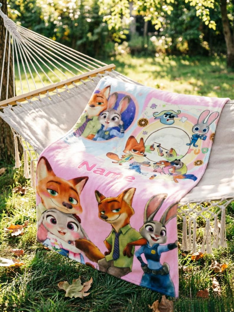 May include: A soft pink fleece blanket featuring colorful cartoon illustrations of a fox and a rabbit character from Zootopia. The design includes multiple scenes of the characters, a circular frame, and the word Name printed in a bold pink font.