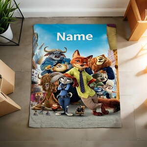 May include: A rectangular blanket featuring characters from the animated film Zootopia. The blanket has a blue sky background with the word "Name" in white. The characters include a fox, a bunny, a lion, and other animals.