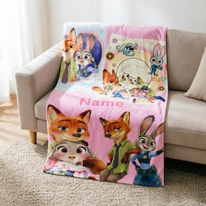 May include: A soft pink fleece blanket featuring Zootopia characters Nick Wilde and Judy Hopps in various poses. The design includes a circular graphic with smaller character illustrations, the word Name in pink text, and a repeating pattern.