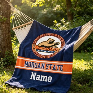 Könnte beinhalten: Eine marineblaue Decke mit den Wörtern "MORGAN STATE" und "Name" in Weiß. Die Decke hat einen orangefarbenen Streifen und ein kreisförmiges Logo mit einem Bären. Die Decke liegt über einer Hängematte.