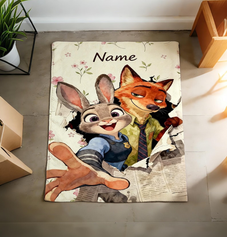 May include: A rectangular blanket featuring a cartoon rabbit and fox character illustration. The design includes a floral background, the word Name at the top, and a newspaper print effect where the characters appear to be tearing through the paper.