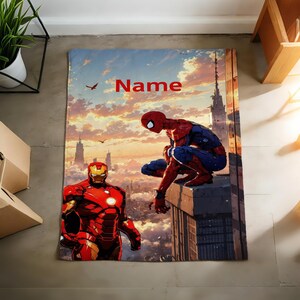 May include: A rectangular blanket featuring a printed graphic of Spider-Man crouching on a building ledge and Iron Man standing in the foreground. The background depicts a city skyline at sunset with clouds and birds. The word Name is printed in red.