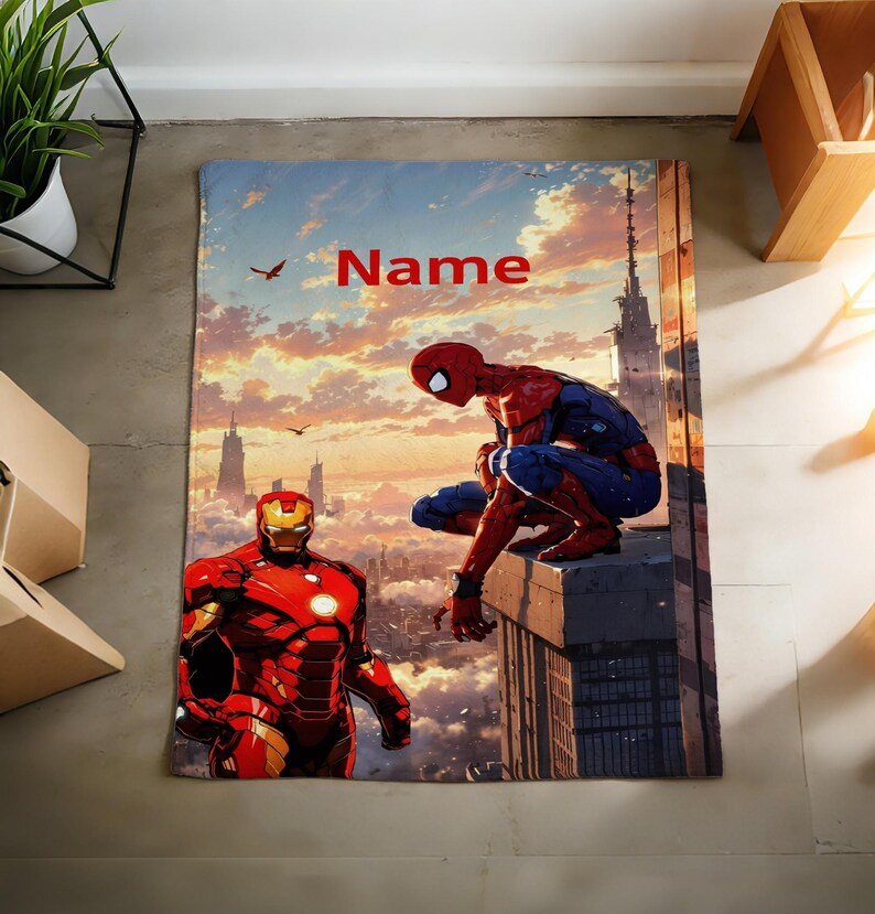 May include: A rectangular blanket featuring a printed graphic of Spider-Man crouching on a building ledge and Iron Man standing in the foreground. The background depicts a city skyline at sunset with clouds and birds. The word Name is printed in red.