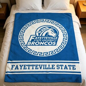 Puede incluir: Una manta de forro polar azul con el logotipo blanco de los Fayetteville State University Broncos, encerrado en un borde circular de greca. El texto FAYETTEVILLE STATE UNIVERSITY BRONCOS aparece en el centro, con FAYETTEVILLE STATE debajo.