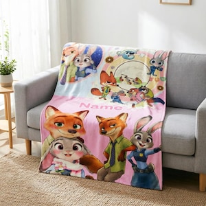 May include: A soft fleece throw blanket featuring Zootopia characters including Nick Wilde and Judy Hopps. The design includes colorful illustrations of the fox and rabbit, a circular scene, and the word Name printed in pink on a light pink background.
