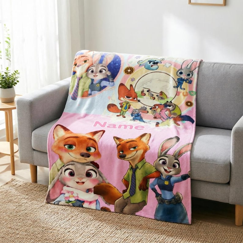 May include: A soft fleece throw blanket featuring Zootopia characters including Nick Wilde and Judy Hopps. The design includes colorful illustrations of the fox and rabbit, a circular scene, and the word Name printed in pink on a light pink background.