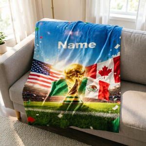 Può includere: Una morbida coperta con un trofeo di calcio dorato al centro, circondato dalle bandiere degli Stati Uniti, del Canada e del Messico. Il design presenta uno sfondo da stadio, coriandoli e la parola Name stampata in bianco in alto.