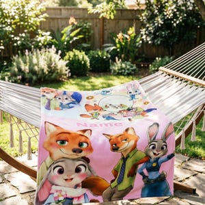May include: A soft pink fleece blanket featuring a colorful graphic print of characters from Zootopia, including Nick Wilde and Judy Hopps. The design includes multiple scenes of the characters and the word Name printed in a light pink font.