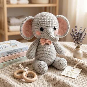 May include: A gray crocheted elephant toy with large ears featuring pink inner sections, black safety eyes, pink cheeks, and a small peach-colored bow tie. The elephant is positioned next to a stack of three books and two wooden rings.