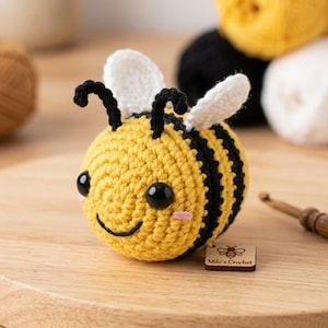 Peut inclure: Un jouet d'abeille crocheté à la main. L'abeille a un corps jaune avec des rayures noires, des ailes blanches et des antennes noires. Elle a un visage souriant avec des joues roses. Une étiquette en bois indique "Milo's Crochet".