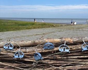 May include: A string of light blue decorative ornaments shaped like small vintage campers, connected by a thin wire and draped across a stack of dry, weathered wooden branches against a backdrop of a coastal landscape and blue sky.