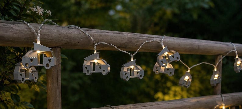 Warmweiß leuchtende LED-Lichterkette mit kleinen Van/Kastenwagen-Motiven als Camping-Deko, dekorativ an einem Holzgeländer im Grünen aufgehängt.