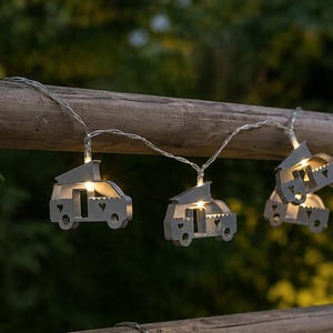 Op de afbeelding: Een slinger van decoratieve lampjes met kleine, grijze campervormen. Elke camper heeft een uitgesneden deur en ramen, met een warm, gloeiend licht van binnen. De lampjes zijn geregen aan een dunne draad, perfect voor buitendecoratie.