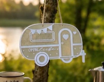 May include: A beige and white hanging sign shaped like a vintage camper trailer. The sign features a white outline, a heart on the door, bunting flags, and the text "CAMPING Liebe" in white script. It hangs from a light-colored string.