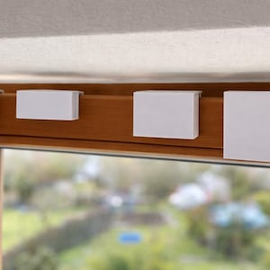 Op de afbeelding: Vier witte rechthoekige raamblindclips zijn bevestigd aan een houten raamkozijn. De clips zijn gelijkmatig verdeeld en lijken gemaakt van een glad, mat materiaal. De achtergrond toont een wazig beeld van een groene buitenscène.