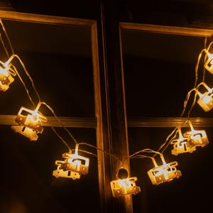 Op de afbeelding: Een reeks warmwitte decoratieve lampjes in de vorm van kleine kampeerwagens. De lampjes zijn gespannen over een houten raamkozijn, waardoor een gezellige en uitnodigende sfeer ontstaat. De lampjes lichten op tegen een donkere achtergrond.