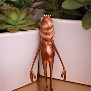 Può includere: Statuetta color bronzo di una creatura con testa bulbosa, braccia lunghe e gambe sottili. La statuetta ha piccoli occhi neri ed è in posa con le braccia tese. Lo sfondo comprende fioriere bianche e piante verdi.