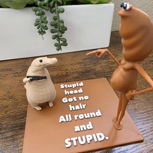 May include: A brown and beige 3D-printed figurine set. One figure resembles a creature with a long neck, pointing at a second figure, a duck-like creature holding a black object. A brown base displays the text "Stupid head Got no hair All round and STUPID."